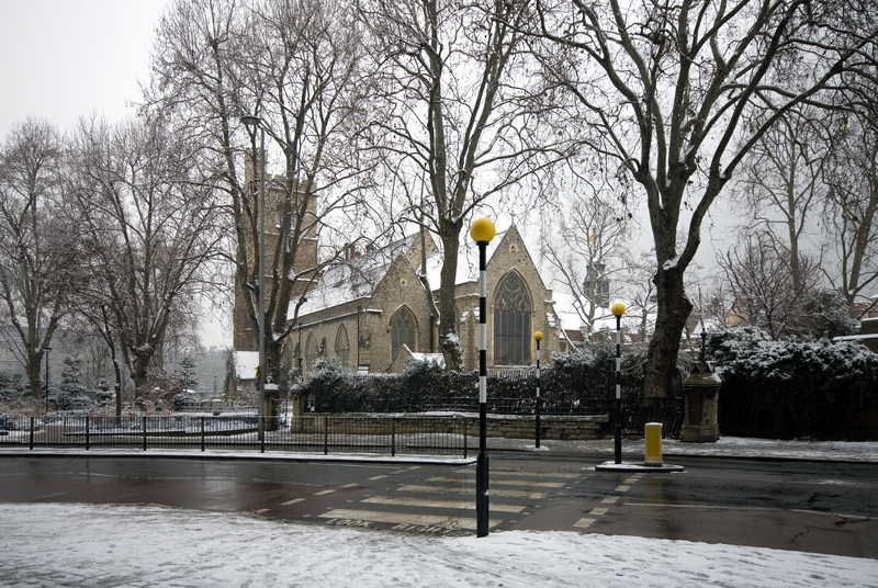 A historic stone church with pointed Gothic-style windows and steeply pitched roofs is situated behind a black metal fence in a small outdoor space. The church is surrounded by leafless trees and low hedges, all partially covered in snow, indicating a winter scene. In the foreground, there is a pedestrian crossing with white-striped markings on the wet asphalt, and a series of yellow and black bollards and street lamps lining the street. The area appears quiet and well-maintained, with the snow creating a natural contrast against the dark pavement and the stone walls of the church. The overall scene suggests a tranquil moment in a London suburb during winter, with landscaping elements and outdoor maintenance in view, fitting naturally with gardening and outdoor service themes.