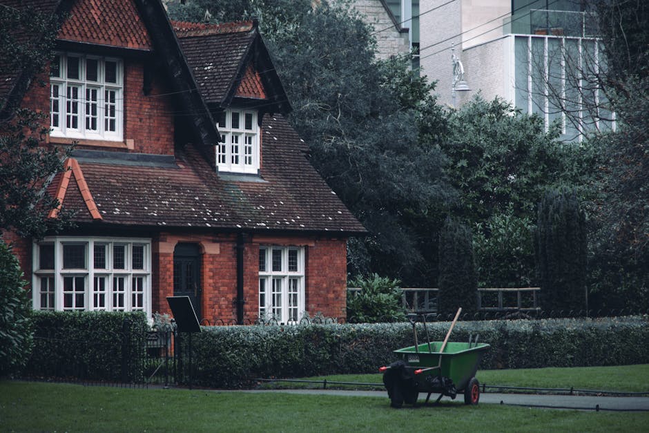 A picturesque front yard of a red brick house features a well-maintained lawn bordered by neatly trimmed hedges and a low fence. The house has white-framed windows with multiple panes, and a dark door