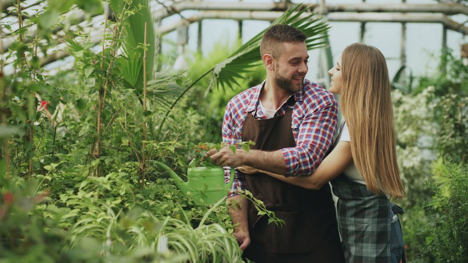 A young woman in a white t-shirt, ripped jeans, and a checked apron holds a rake while standing in a lush garden, smiling at a man dressed in a plaid shirt, dark apron, and trousers who is holding a p